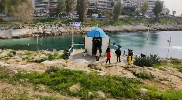 Mała kapliczka nad wodą, z grecką flagą, na tle budynków. Widok na pireus plaża.