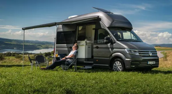 Mężczyzna odpoczywa na krześle obok szarego VW Transporter kampera, rozkoszując się widokiem jeziora i wzgórz.