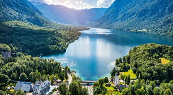 Malownicze jezioro Bohinj otoczone zielonymi wzgórzami i górami. Widać most, kościół i hotel.