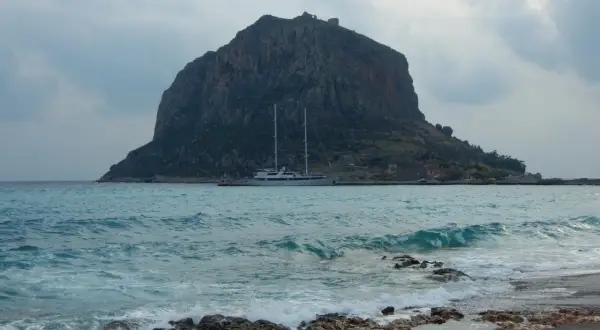 Monemvasia, Grecja: skalista góra z ruinami zamku na szczycie, jacht na turkusowym morzu i kamienista plaża.
