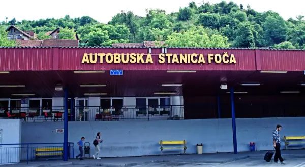 Autobuska stanica Foča, zalesione wzgórza w tle. Mężczyzna z walizką na kółkach, dzieci idące obok. Marka konwertybilna.