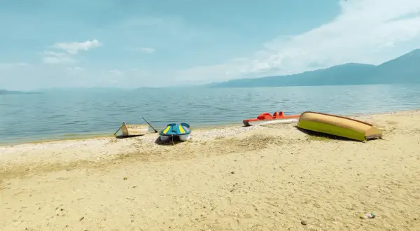 Spokojny brzeg Jeziora Ochrydzkiego w Albanii. Kilka łódek, w tym kolorowa łódka i czerwona rowerowa, odpoczywa na piaszczystej plaży.