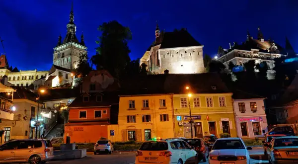 Nocne, oświetlone budynki i wieża zegarowa w Sighișoara. Atrakcje miasta nocą, z samochodami na pierwszym planie.