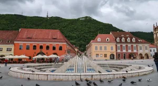 Fontanna na placu w Braszowie, z kolorowymi kamienicami i napisem "BRASOV" na wzgórzu. Wiele gołębi spaceruje wokół.