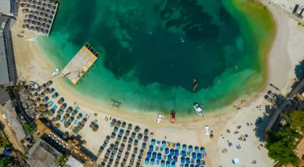 Widok z lotu ptaka na turkusowe morze i piaszczystą plażę z rzędami leżaków i parasoli. Woda jest krystalicznie czysta, widać ludzi pływających i łodzie. To idealne miejsce na relaks na **tirana plaże**.