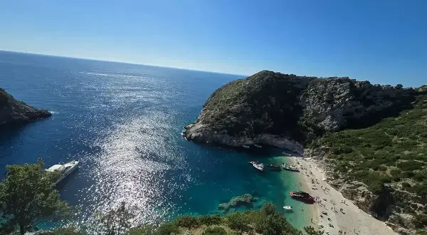 Słoneczna zatoka grama z turkusową wodą, kamienistą plażą pełną ludzi i łodziami przycumowanymi do brzegu.