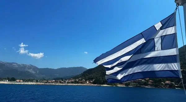 Flaga Grecji powiewa nad błękitnym morzem, w tle widać górzyste wybrzeże z zabudowaniami. Idealny dzień na plażowanie w Grecji.