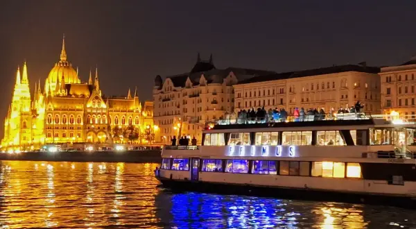 Nocny rejs po Dunaju w Budapeszcie. Oświetlony Parlament i statek z pasażerami na pokładzie odbijają się w wodzie.
