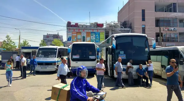 Autobusy na dworcu, jeden z nich jedzie do Vlory, inny do Tirany. Ludzie czekają na podróż.