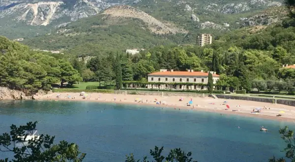 Malownicza zatoka z plażą w Czarnogórze. Woda jest turkusowa, a plaża piaszczysta, z kilkoma osobami odpoczywającymi pod parasolami. W tle widać zielone wzgórza i elegancki budynek.