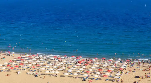 Słoneczny brzeg tętni życiem. Tłumy plażowiczów odpoczywają pod parasolami, kąpią się w morzu i opalają na piasku.