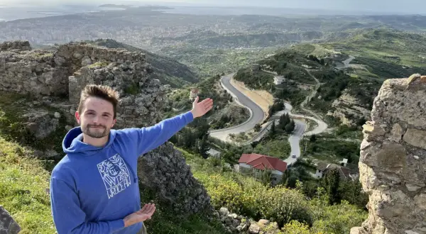 Mężczyzna na tle malowniczego krajobrazu z krętą drogą i miastem w oddali. To piękna Albania zimą, choć zieleń sugeruje wczesną wiosnę.
