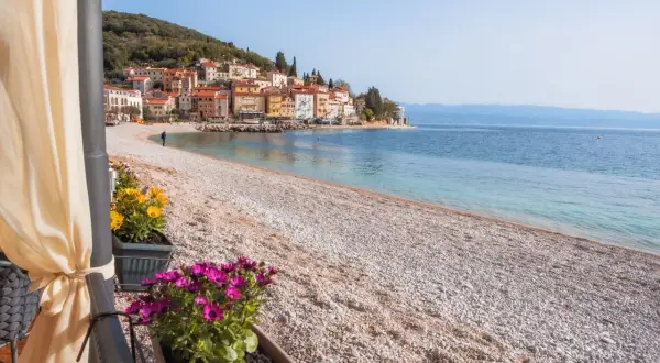 Kamienista plaża z widokiem na kolorowe domki na wzgórzu. To właśnie takie widoki sprawiają, że Istria ma najpiękniejsze plaże.