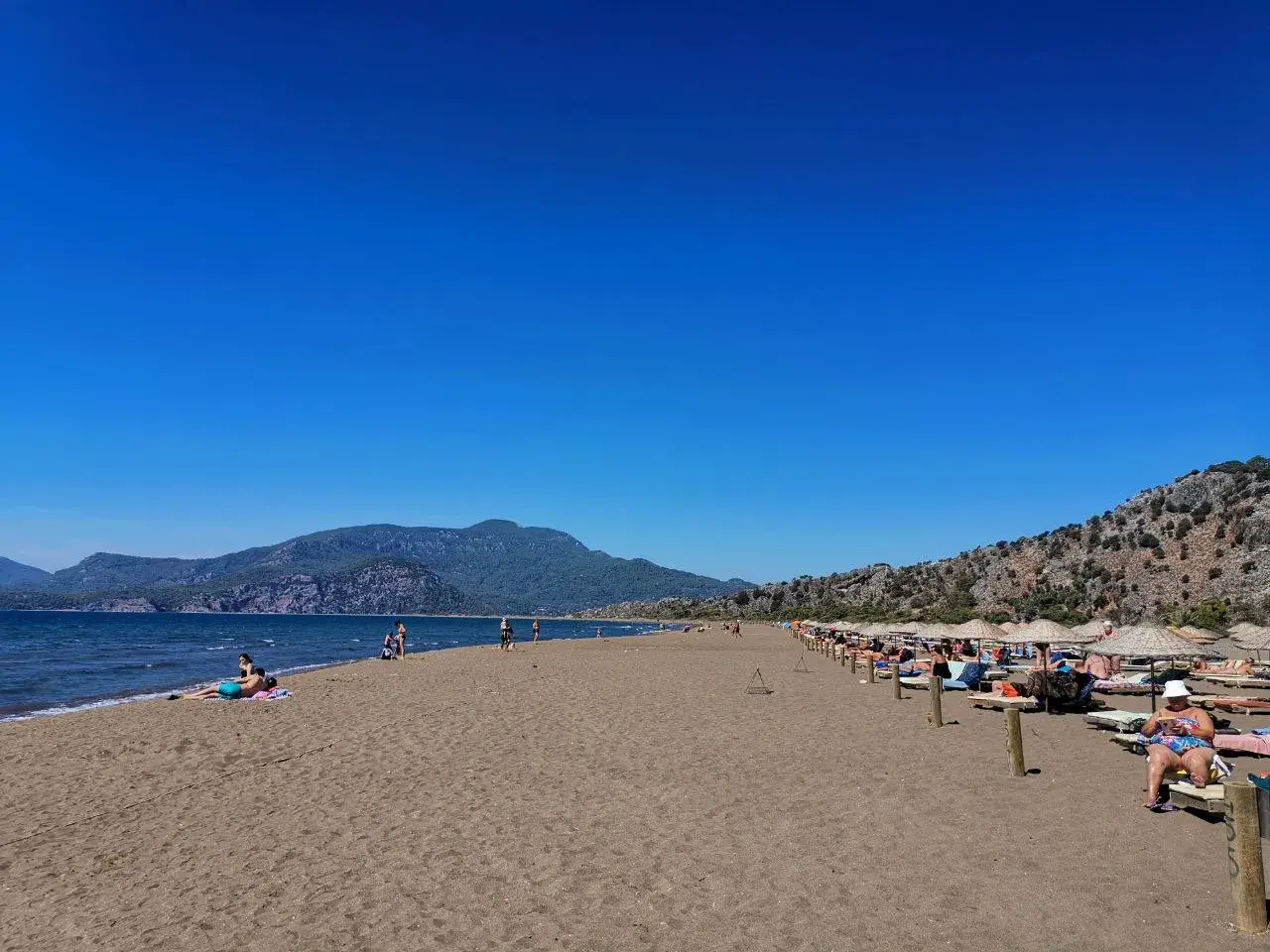 Plaża żółwi w Turcji, z turkusowym morzem i górami w tle. Ludzie odpoczywają na leżakach pod parasolami.