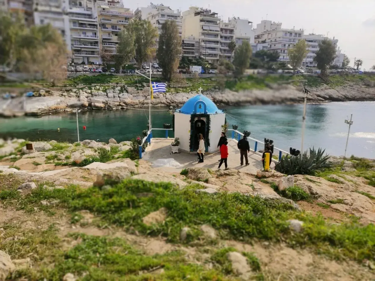 Mała kapliczka nad wodą, z grecką flagą, na tle budynków. Widok na pireus plaża.