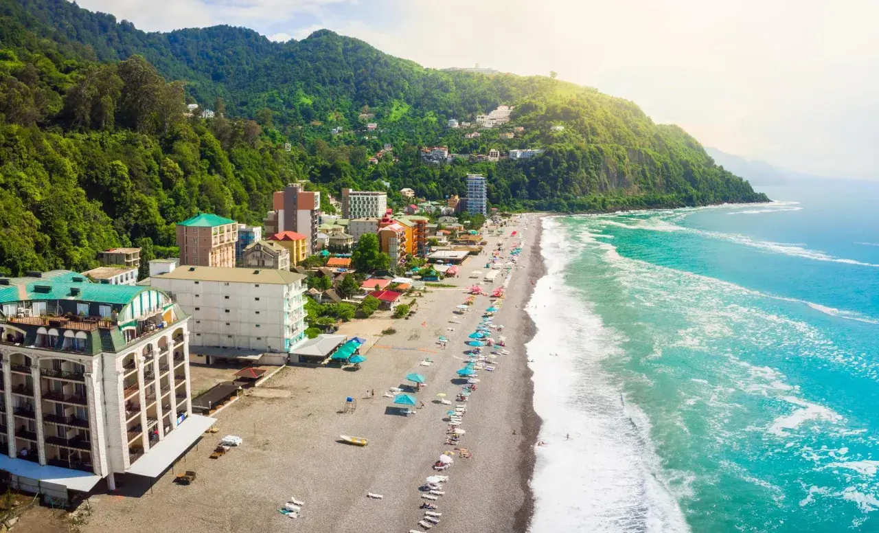 Widok na plażę i hotele w Batumi, Gruzja. Zielone wzgórza schodzą do turkusowego morza, a na kamienistej plaży widać parasole i leżaki.