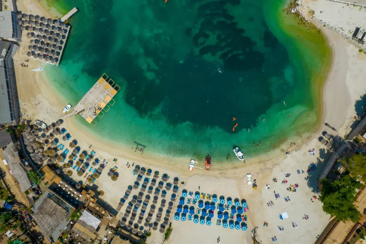 Widok z lotu ptaka na turkusowe morze i piaszczystą plażę z rzędami leżaków i parasoli. Woda jest krystalicznie czysta, widać ludzi pływających i łodzie. To idealne miejsce na relaks na **tirana plaże**.
