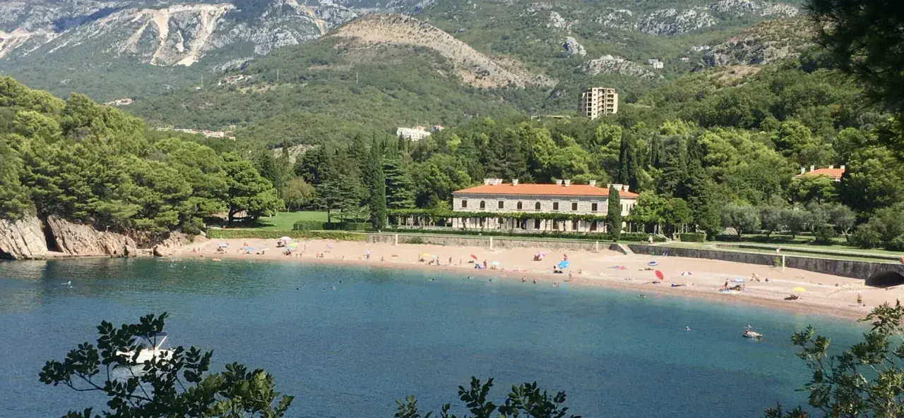 Malownicza zatoka z plażą w Czarnogórze. Woda jest turkusowa, a plaża piaszczysta, z kilkoma osobami odpoczywającymi pod parasolami. W tle widać zielone wzgórza i elegancki budynek.