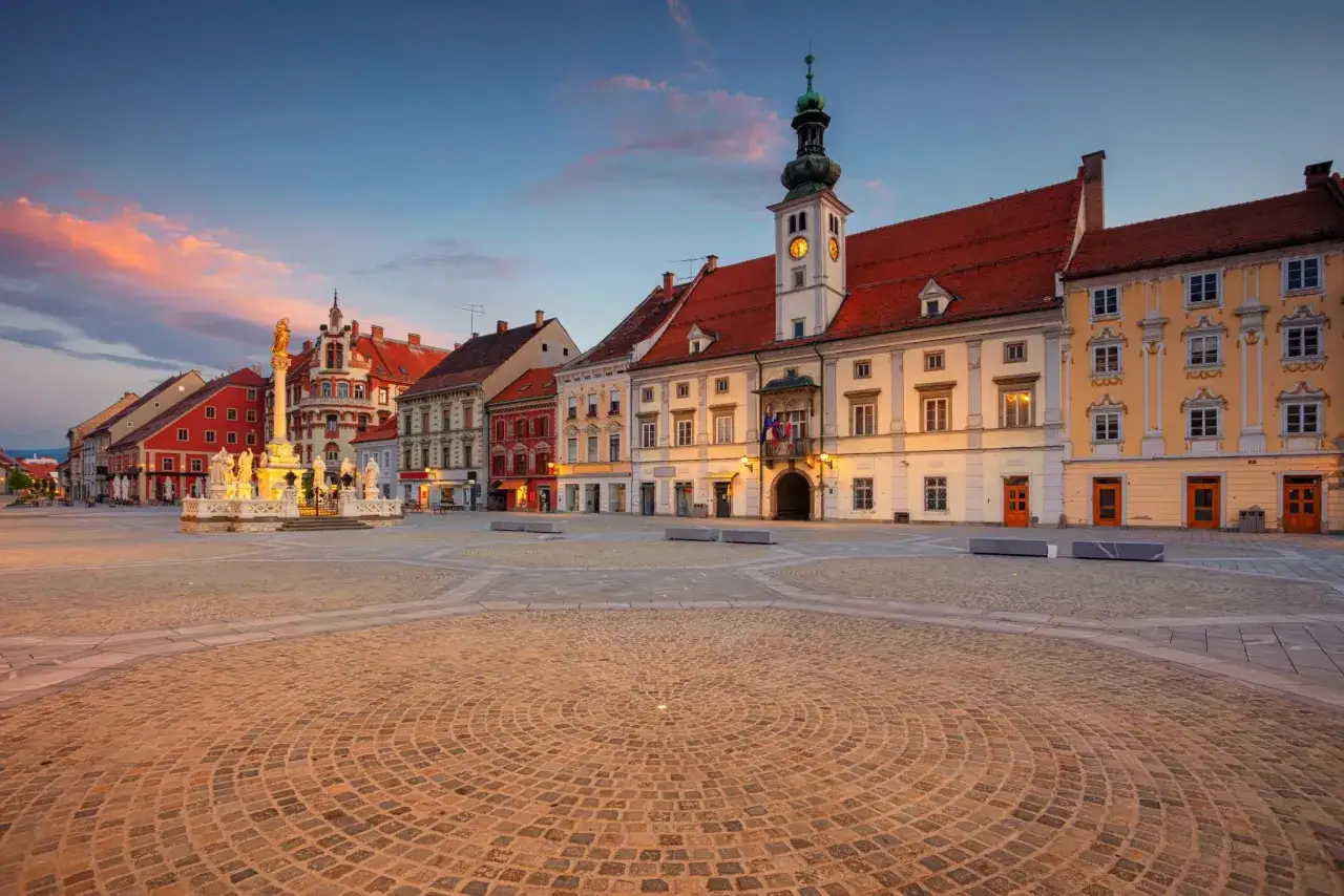 Rynek w Mariborze, Słowenia, o zachodzie słońca. Kolorowe chmury nad zabytkowymi kamienicami i fontanną.