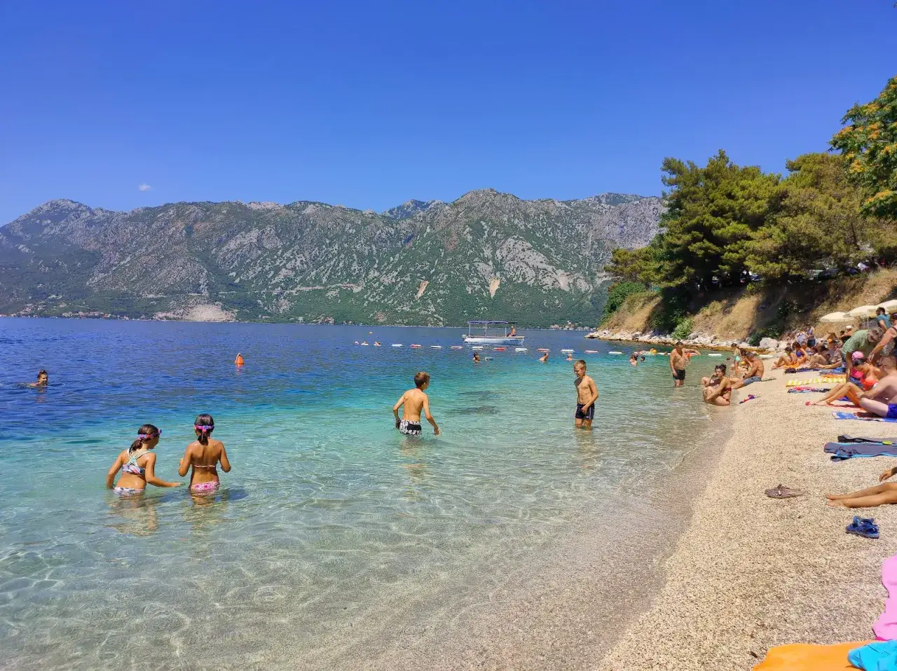 Słoneczny dzień na Perast beach. Ludzie kąpią się w krystalicznie czystej wodzie, podczas gdy w tle widać góry i łódź.