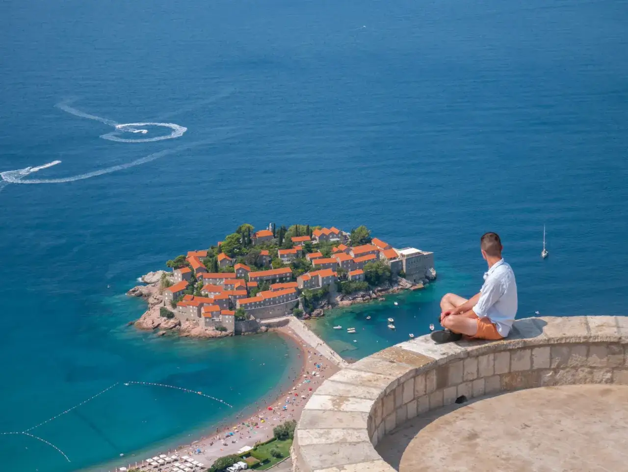 Mężczyzna siedzi na murze, podziwiając wyspę Sveti Stefan. Czy Czarnogóra jest bezpieczna? Tak, wygląda na spokojną i malowniczą.