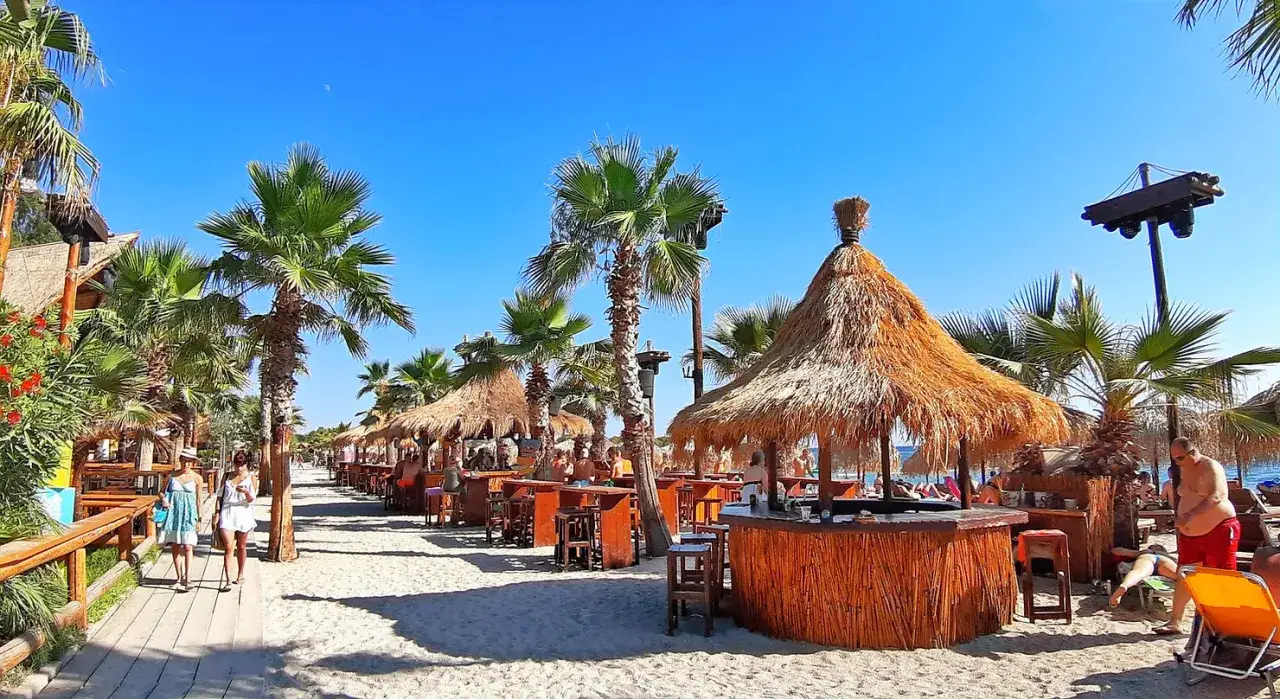 Słoneczny dzień na plaży w Atenach. Palmy, drewniane bary z krytymi strzechą dachami i piasek tworzą wakacyjny klimat.