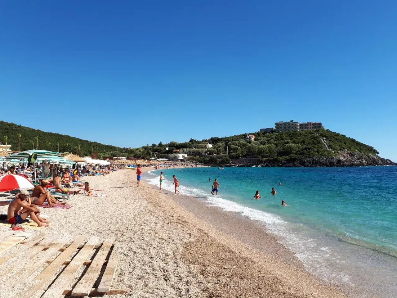 Słoneczny dzień na plaży Jale, z turystami odpoczywającymi pod parasolami i kąpiącymi się w turkusowym morzu.