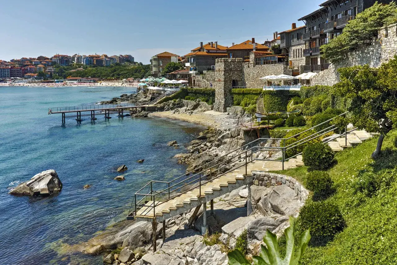 Kamieniste wybrzeże z kamiennymi schodami prowadzącymi do morza, w tle plaża i zabudowania. Zastanawiasz się, ile kosztuje lot do Bułgarii?