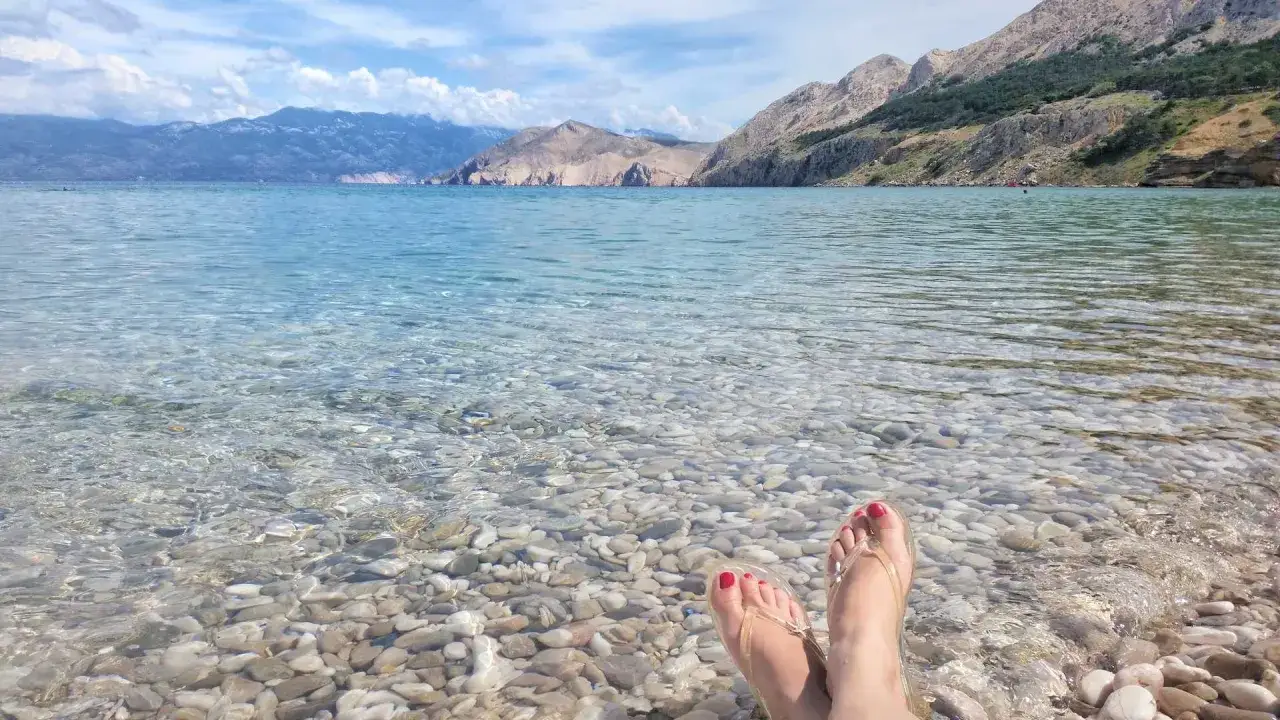 Stopy w klapkach zanurzone w krystalicznie czystej wodzie na kamienistej plaży w Baška, Chorwacja. W tle góry i błękitne niebo.