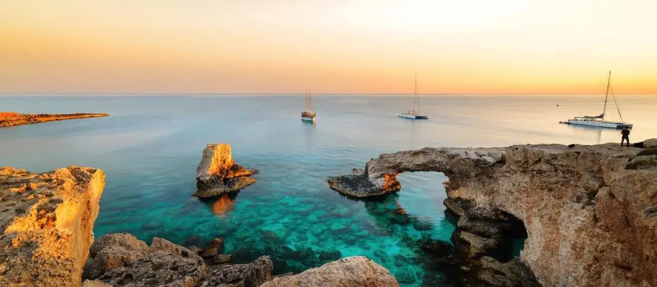 Zachód słońca nad Cape Greco na Cyprze. Turkusowa woda, łuki skalne i żaglówki tworzą malowniczy krajobraz.