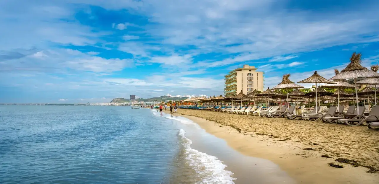 Plaża w jednym z albańskich miast, z rzędem leżaków i parasoli. W tle widać morze i zabudowania.