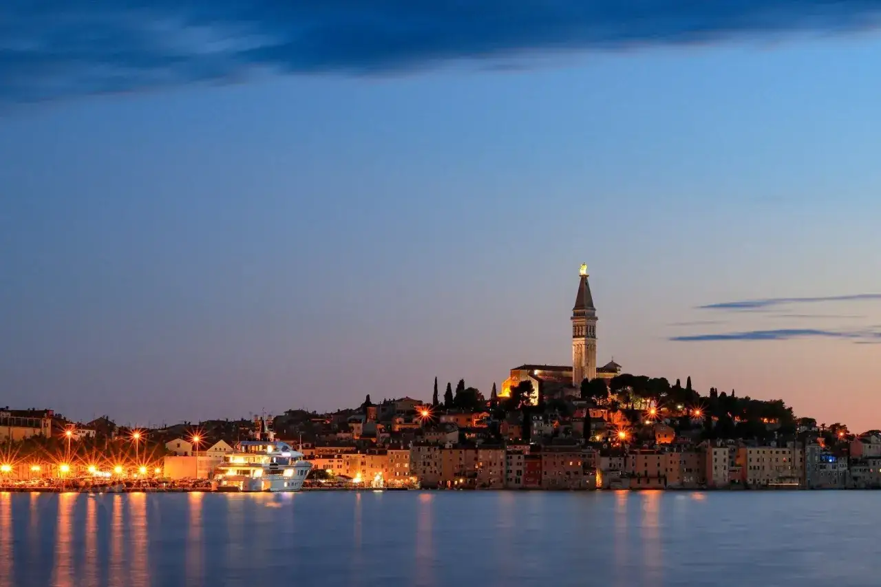 Wieczorna panorama Chorwacji, Istria atrakcje: miasto oświetlone blaskiem latarni, z okazałym jachtem i wieżą kościelną górującą nad zatoką.