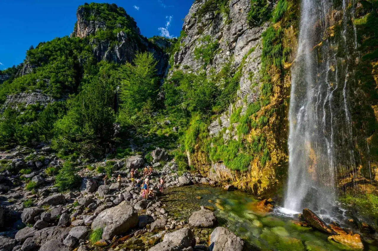 Wodospad Grunas w Theth, Albania, kaskada spływa po skalnej ścianie porośniętej zielenią, u podnóża ludzie odpoczywają nad krystalicznie czystą wodą.