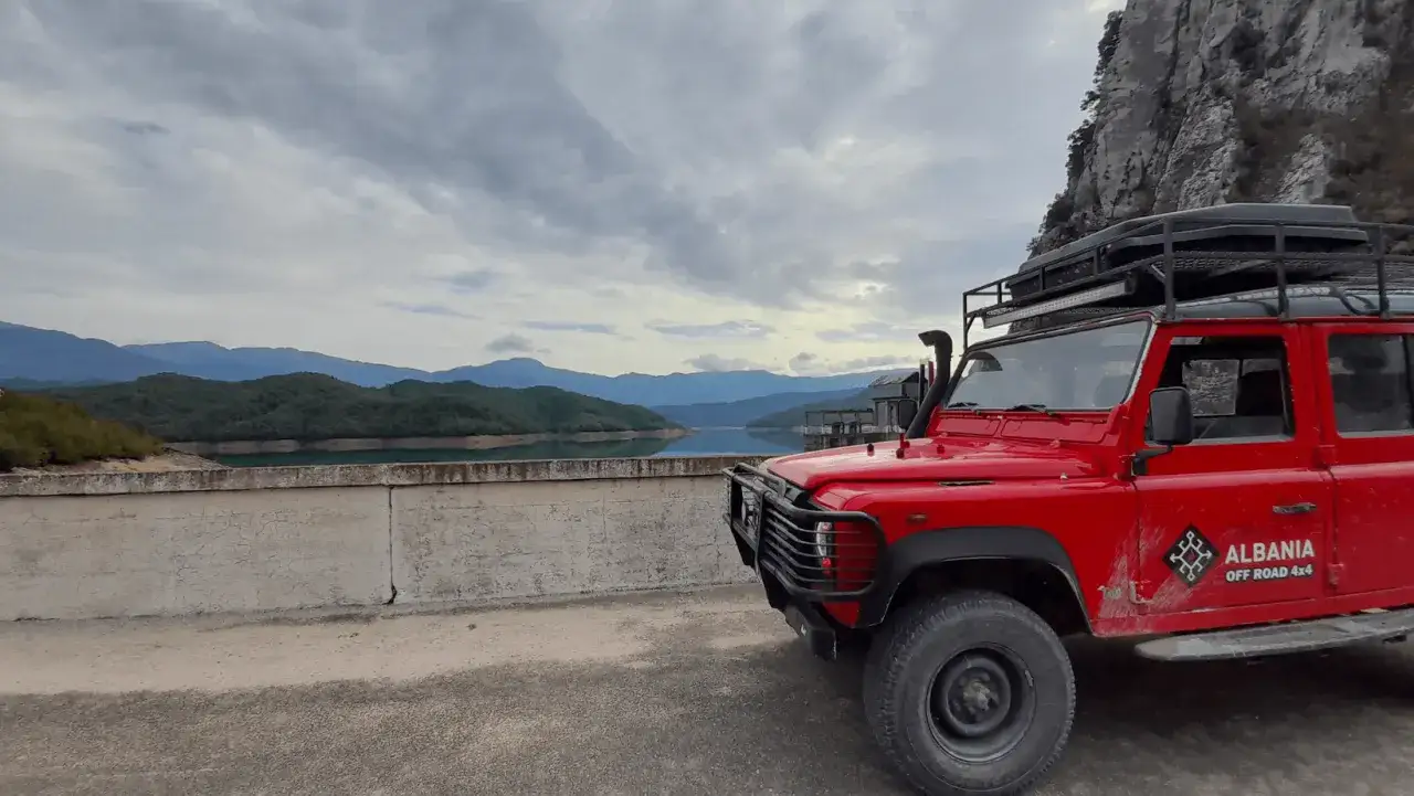 Czerwony Land Rover na tle g&oacute;rskiego jeziora. Chmurne niebo, typowa albania pogoda styczeń.