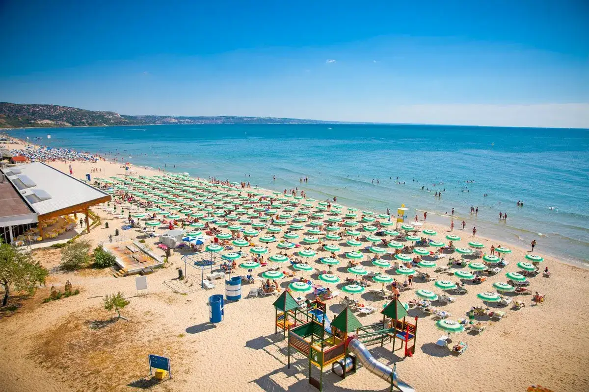 Słoneczna plaża w Bułgarii, pełna zielonych parasoli i wypoczywających ludzi. Zastanawiasz się, ile kosztuje lot do Bułgarii?