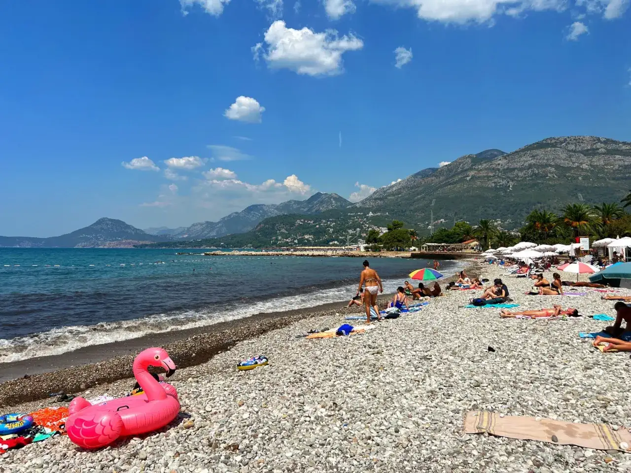 Sutomore atrakcje: plaża z kamykami, morze, góry w tle i mnóstwo ludzi odpoczywających na ręcznikach. Różowy flaming unosi się na wodzie.
