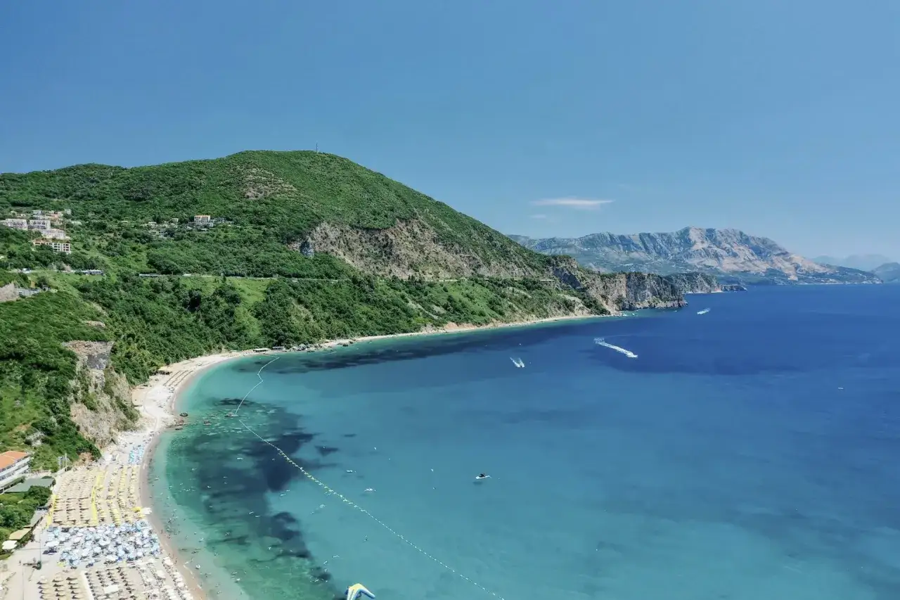 Malownicze budva plaze z turkusową wodą, zielonymi wzgórzami i górami w tle. Widać łodzie i parasole na plaży.