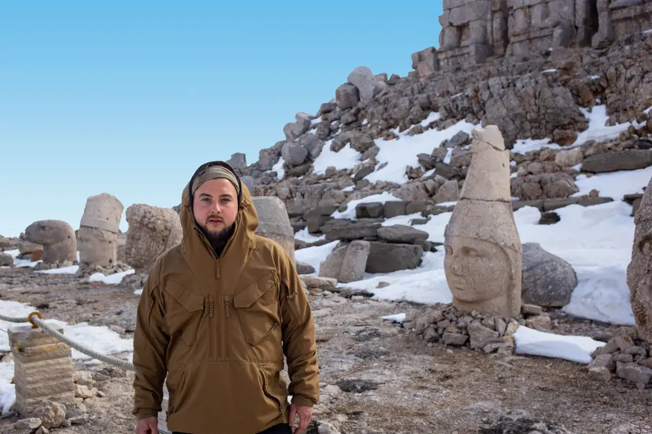 Mężczyzna w brązowej kurtce na tle starożytnych posągów na górze Nemrut. Śnieg i kamienie tworzą surowy krajobraz.