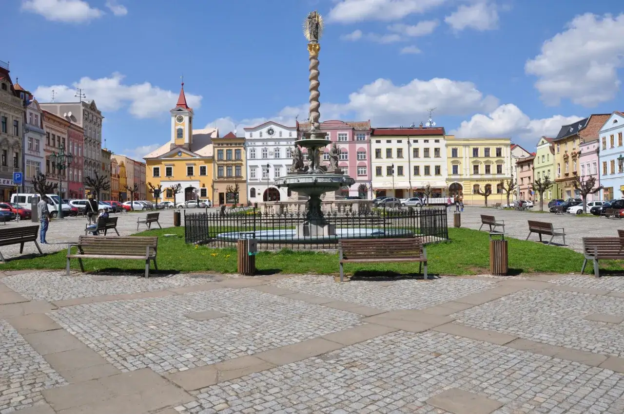 Kolorowy rynek w Broumovie z fontanną i kolumną maryjną. W tle kolorowe kamienice, a na pierwszym planie ławki i brukowana nawierzchnia. Broumov atrakcje zachęcają do spaceru.