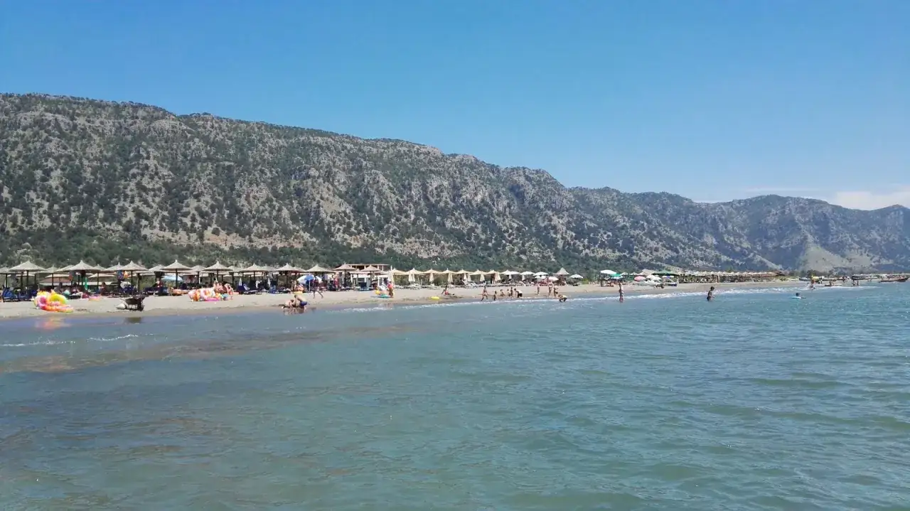 Słoneczny dzień na plaży Velipoja. Ludzie odpoczywają pod parasolami, kąpią się w morzu, a w tle wznoszą się góry.