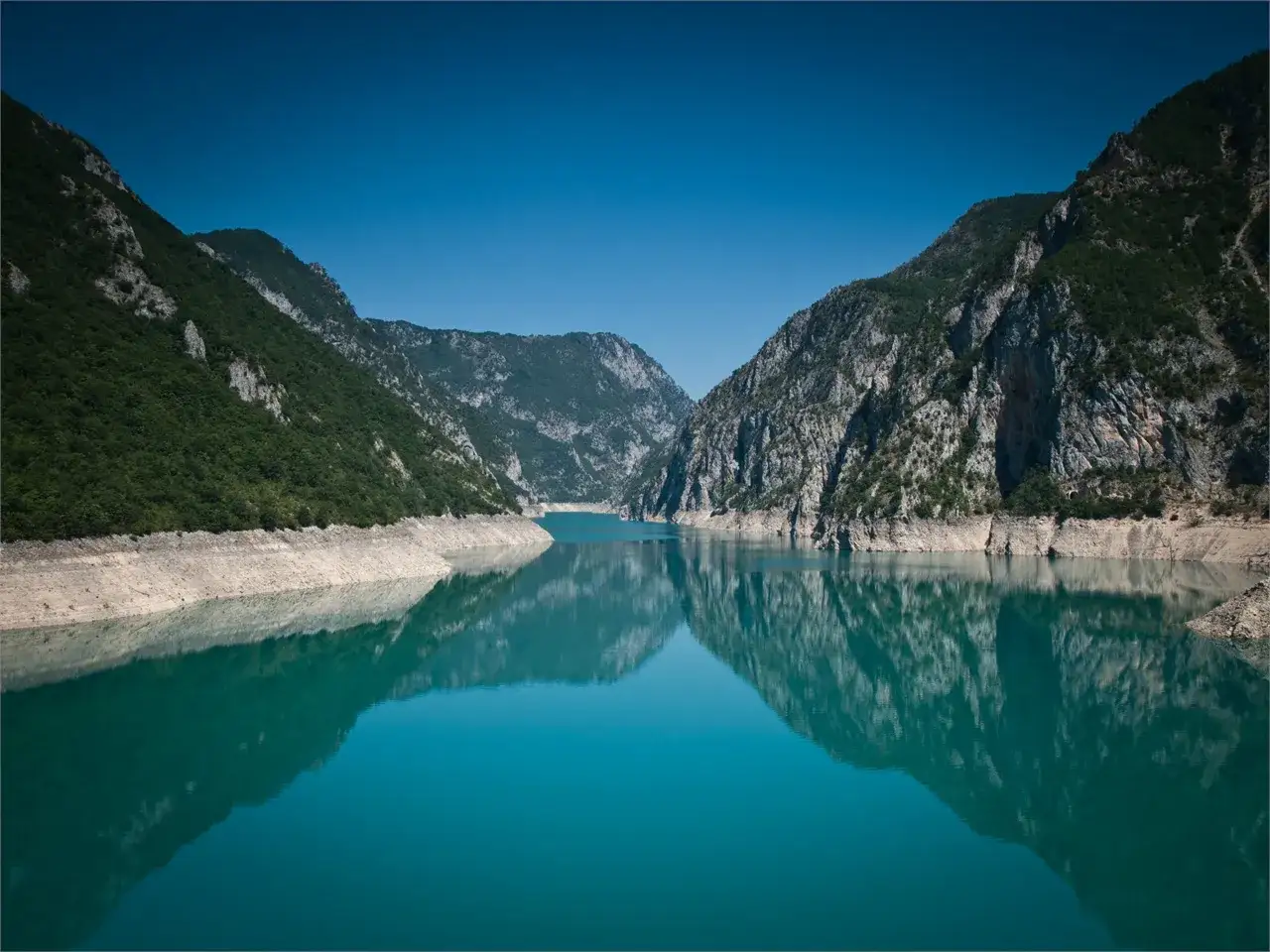 Turkusowe wody Pivsko Jezero odbijają skaliste góry porośnięte zielenią.