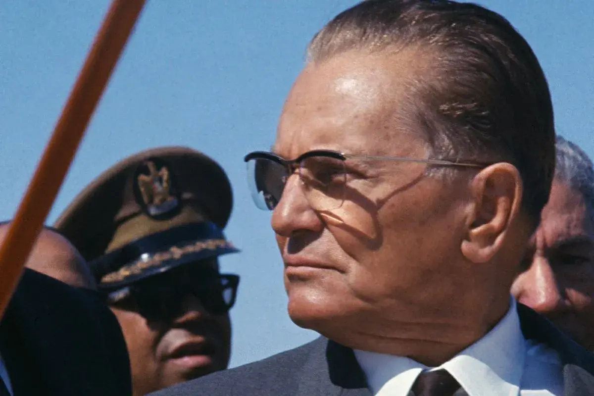 Josip Broz Tito w okularach, w otoczeniu innych mężczyzn, w tym jednego w wojskowym nakryciu głowy.