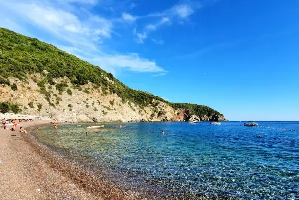 Kamienista plaża z turkusową wodą, zielonymi wzgórzami i kilkoma łodziami na morzu.