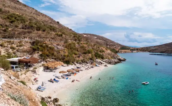 Słoneczna plaża w Porto Palermo z turkusową wodą, leżakami i łódkami.