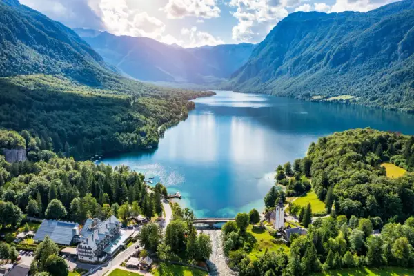 Malownicze jezioro Bohinj otoczone zielonymi wzgórzami i górami. Widać most, kościół i hotel.