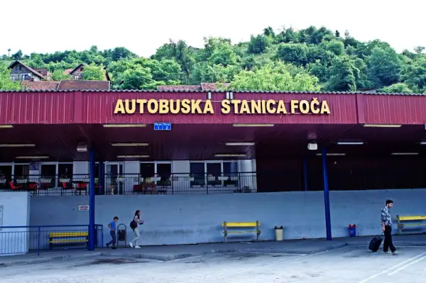 Autobuska stanica Foča, zalesione wzgórza w tle. Mężczyzna z walizką na kółkach, dzieci idące obok. Marka konwertybilna.