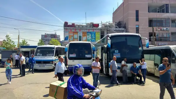 Autobusy na dworcu, jeden z nich jedzie do Vlory, inny do Tirany. Ludzie czekają na podróż.