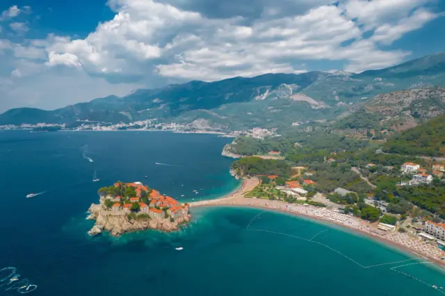 Malownicza czarnogóra wycieczka: wyspa Sveti Stefan połączona z lądem wąskim przesmykiem, turkusowe morze, plaża z leżakami i góry w tle.